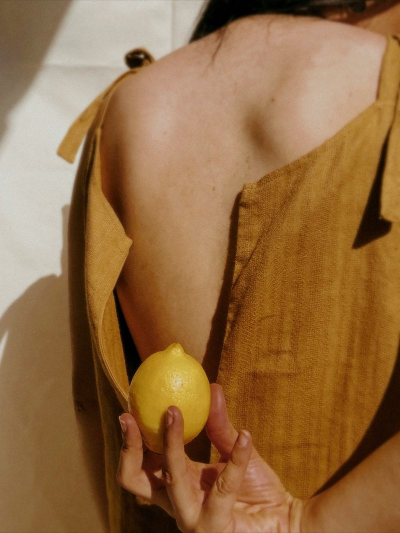 back of a woman in an ochre linen dress, holding a lemon behind her in the sun